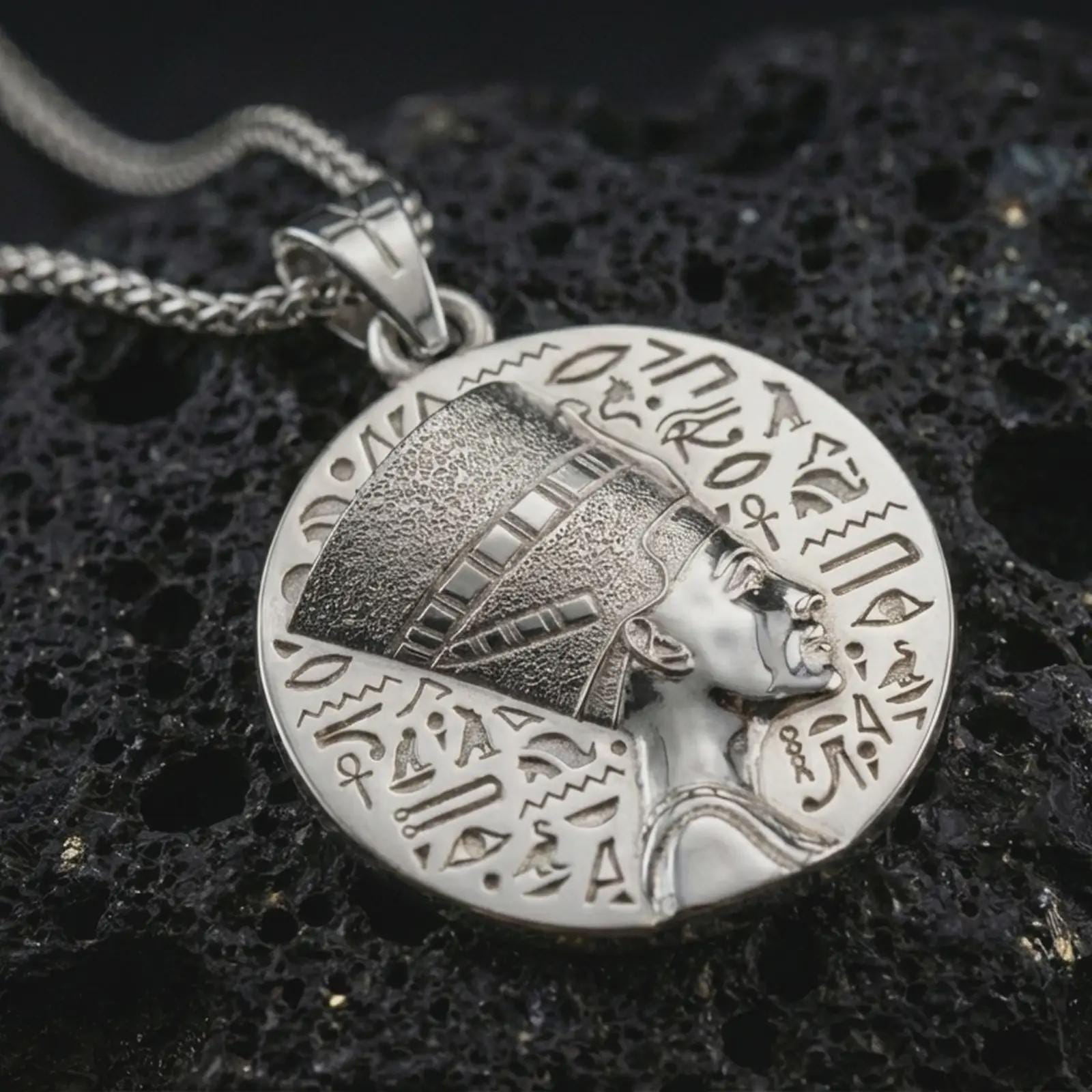 Polished silver Nefertiti medallion pendant necklace displayed on black volcanic rock, shown in a refined macro product shot with Egyptian queen profile, engraved hieroglyphic border, and symbolic bail detail.
