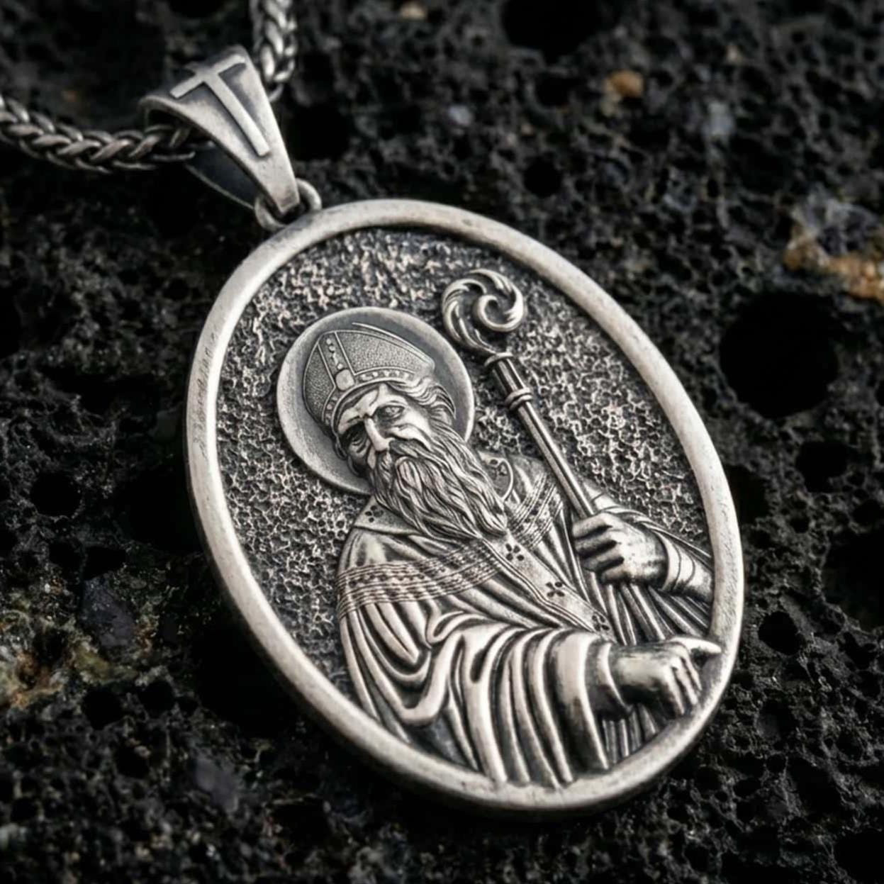 Sterling silver Saint Patrick pendant necklace with an oxidized finish resting on textured volcanic rock, captured in a close-up premium studio composition.