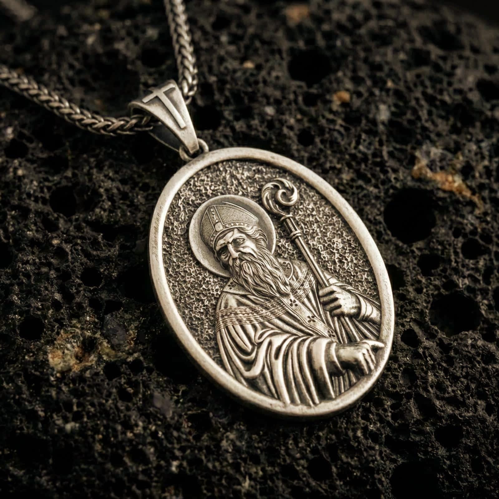 Bronze Saint Patrick pendant necklace with an oxidized finish resting on textured volcanic rock, captured in a close-up premium studio composition.