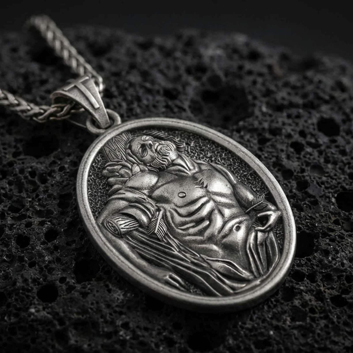 Sterling silver Saint Sebastian pendant necklace with an oxidized finish resting on textured volcanic rock, captured in a close-up premium studio composition.