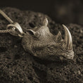 Load image into Gallery viewer, Bronze rhinoceros head pendant necklace with an oxidized finish resting on textured volcanic rock, captured in a close-up premium studio composition.
