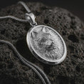 Load image into Gallery viewer, Polished silver snarling wolf pendant necklace displayed on black volcanic rock, shown in a refined macro product shot with fierce canine expression, detailed fur texture, and oval medallion frame.
