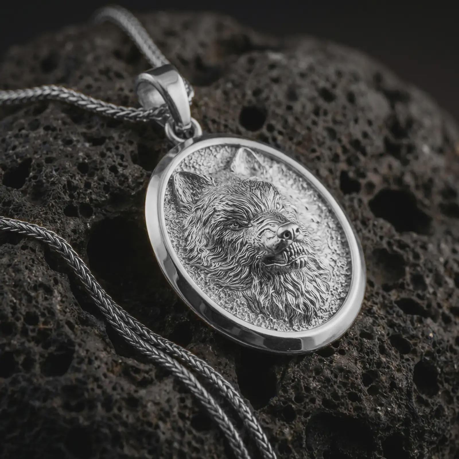 Polished silver snarling wolf pendant necklace displayed on black volcanic rock, shown in a refined macro product shot with fierce canine expression, detailed fur texture, and oval medallion frame.
