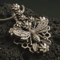 Load image into Gallery viewer, Bronze floral bee pendant necklace with an oxidized finish resting on textured volcanic rock, captured in a close-up premium studio composition with honeycomb, blossoms, and detailed wing relief.
