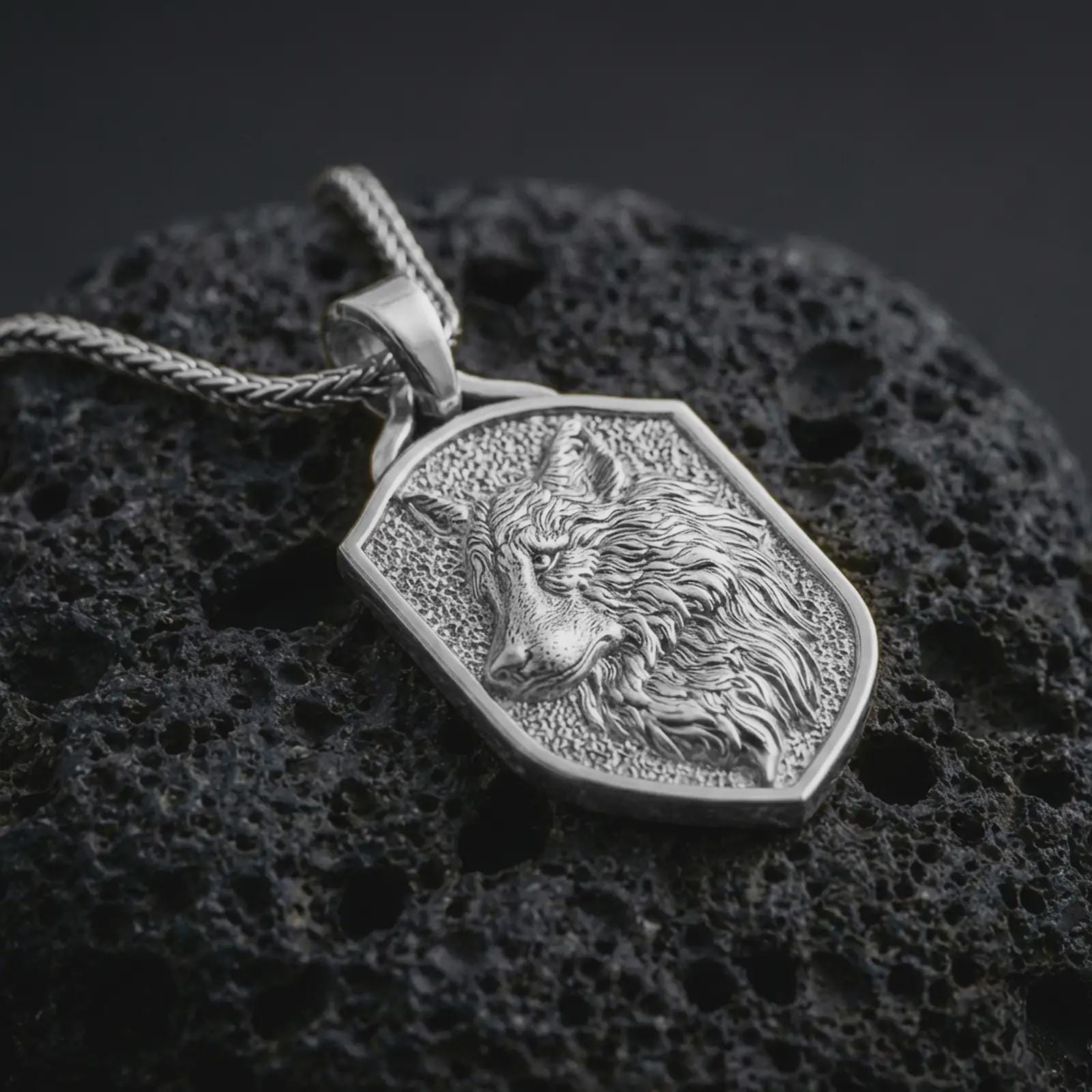 Polished silver wolf shield pendant necklace displayed on black volcanic rock, shown in a refined macro product shot with flowing fur detail, wolf face relief, and geometric shield frame.