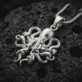 Load image into Gallery viewer, Polished silver octopus pendant necklace displayed on black volcanic rock, shown in a refined macro product shot with curled tentacles, suction cup details, and sculptural nautical relief.
