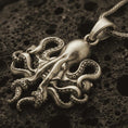 Load image into Gallery viewer, Bronze octopus pendant necklace with an oxidized finish resting on textured volcanic rock, captured in a close-up premium studio composition.

