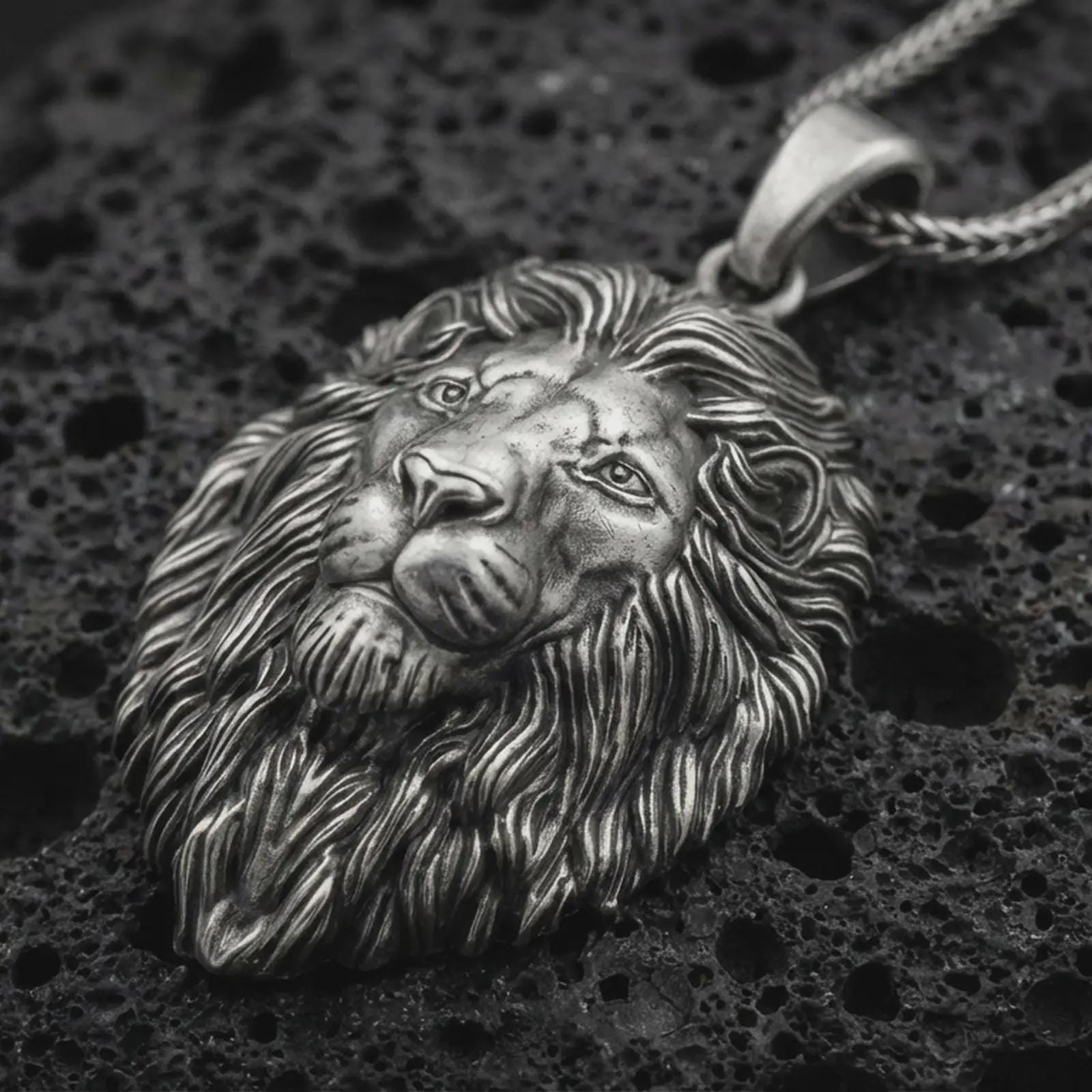 Sterling silver lion head pendant necklace with an oxidized finish resting on textured volcanic rock, captured in a close-up premium studio composition.