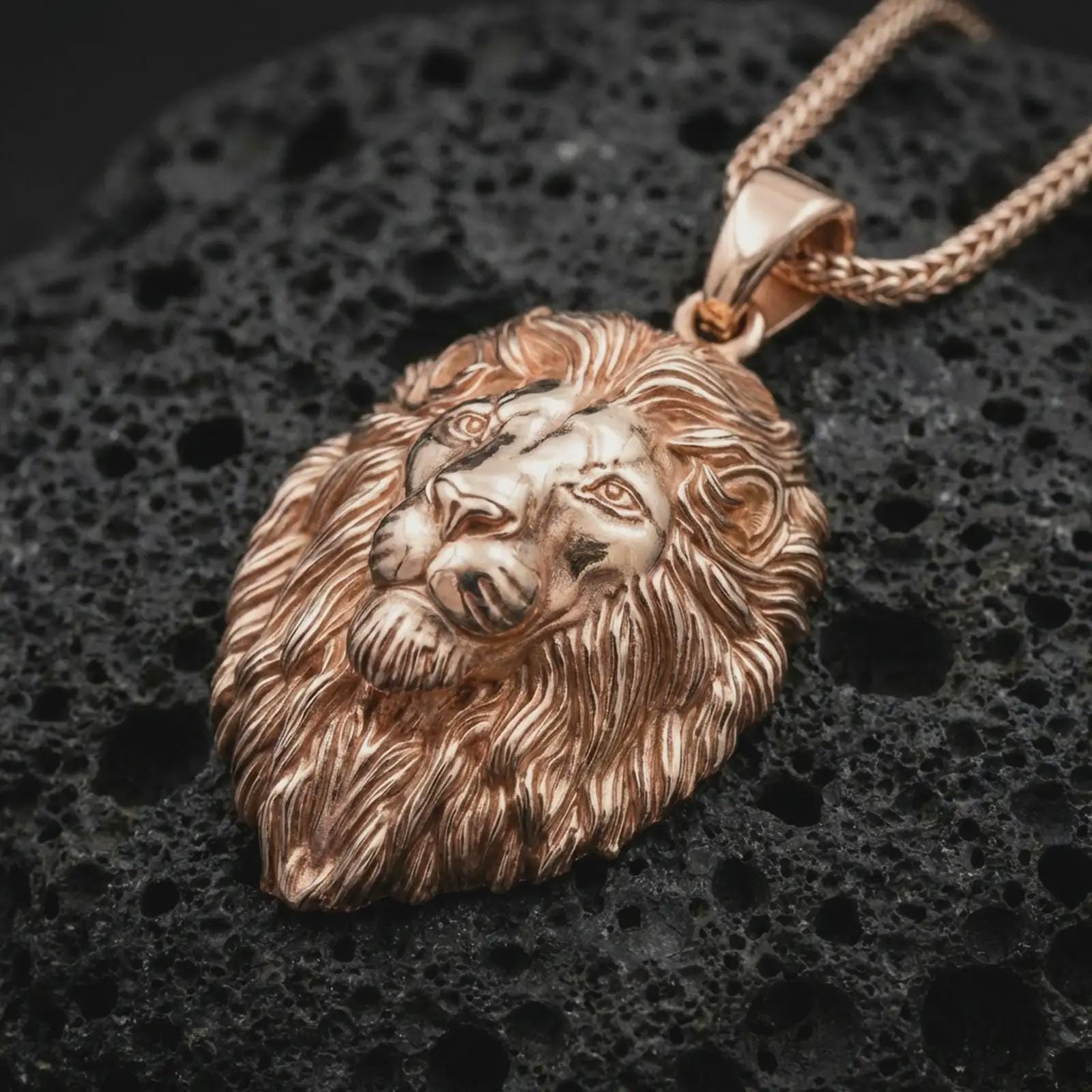 Rose gold plated lion head pendant necklace on dark volcanic rock, photographed in a detailed premium studio macro view.