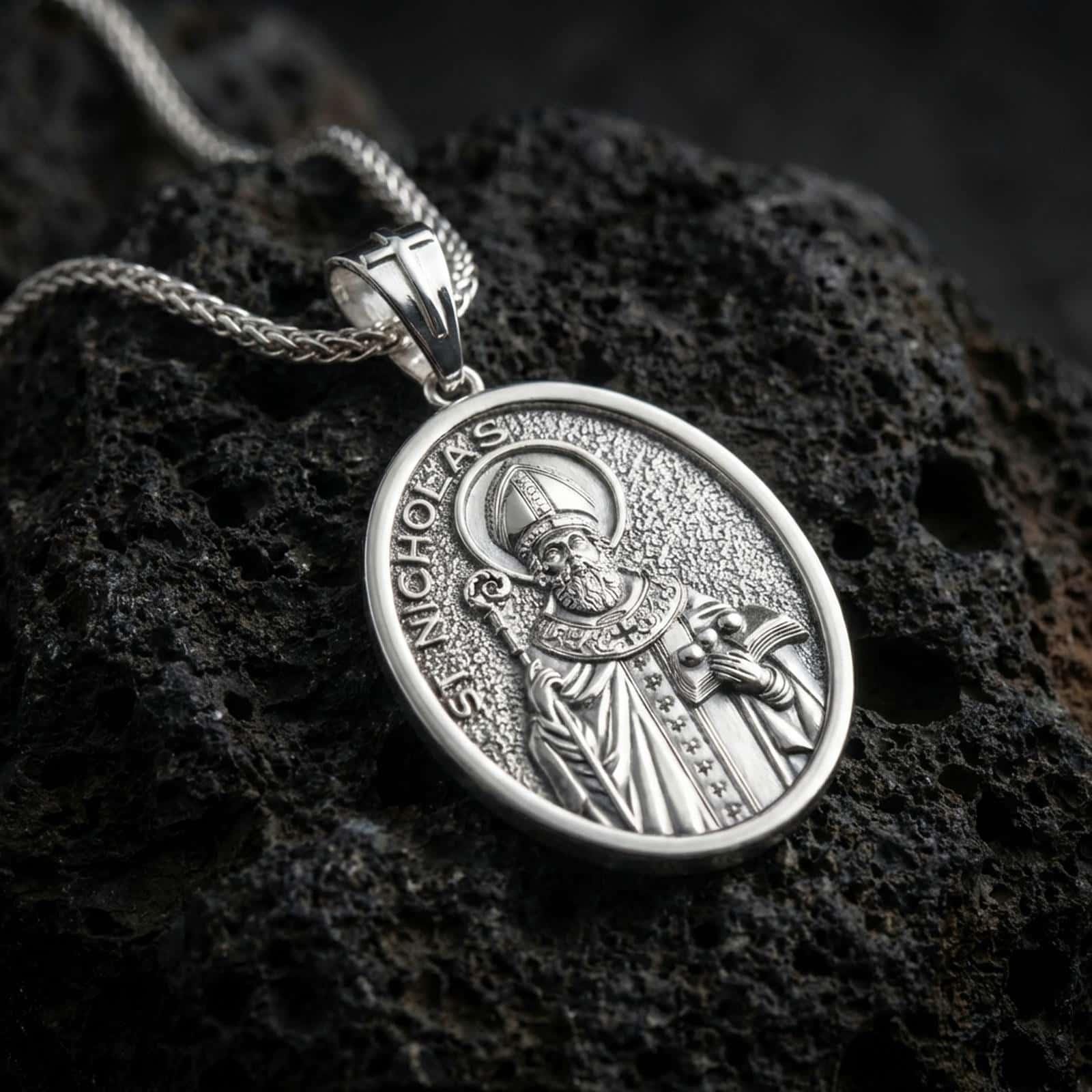 Polished silver Saint Nicholas pendant necklace displayed on black volcanic rock, shown in a refined macro product shot with detailed bishop figure, staff, and engraved lettering.
