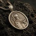 Load image into Gallery viewer, Bronze Saint Nicholas pendant necklace with an oxidized finish resting on textured volcanic rock, captured in a close-up premium studio composition.
