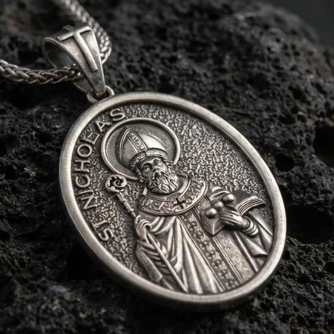 Sterling silver Saint Nicholas pendant necklace with an oxidized finish resting on textured volcanic rock, captured in a close-up premium studio composition.