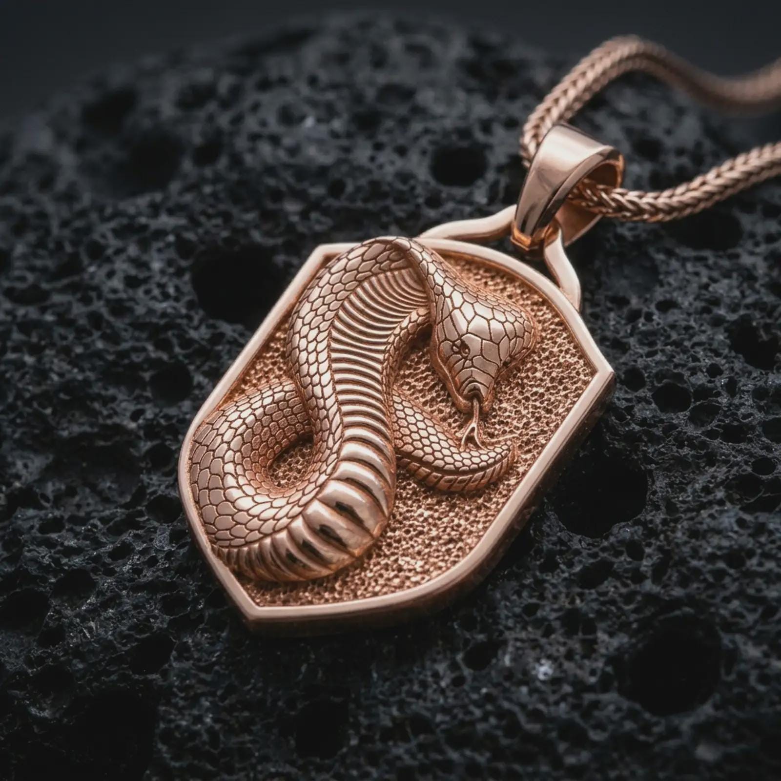 Rose gold plated cobra shield pendant necklace on dark volcanic rock, photographed in a detailed premium studio macro view.