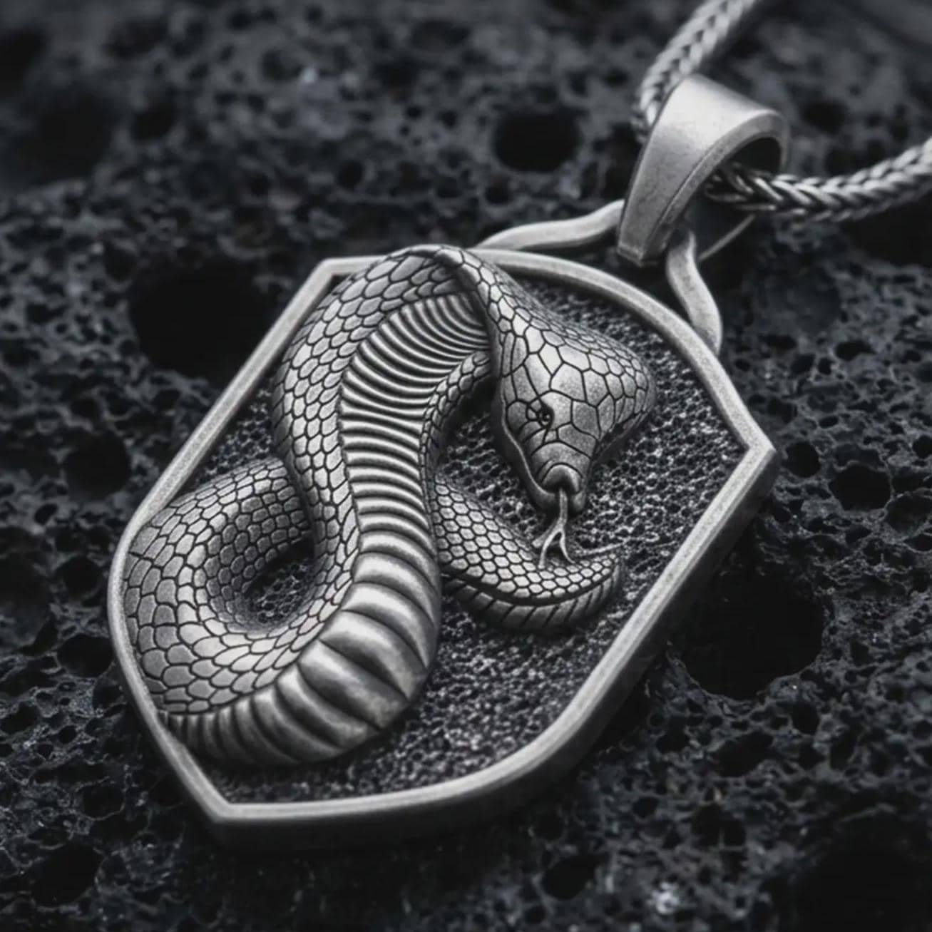Sterling silver cobra shield pendant necklace with an oxidized finish resting on textured volcanic rock, captured in a close-up premium studio composition.