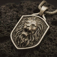 Load image into Gallery viewer, Bronze roaring lion shield pendant necklace with an oxidized finish resting on textured volcanic rock, captured in a close-up premium studio composition.
