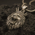 Load image into Gallery viewer, Bronze crowned lion head pendant necklace with an oxidized finish resting on textured volcanic rock, captured in a close-up premium studio composition.
