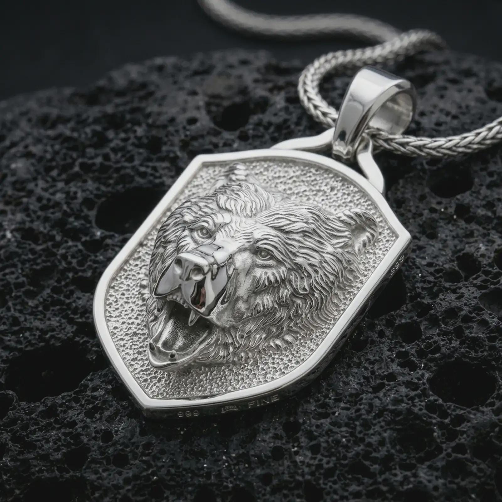 Polished silver roaring bear shield pendant necklace displayed on black volcanic rock, shown in a refined macro product shot with open mouth, detailed fur texture, and angular shield frame.