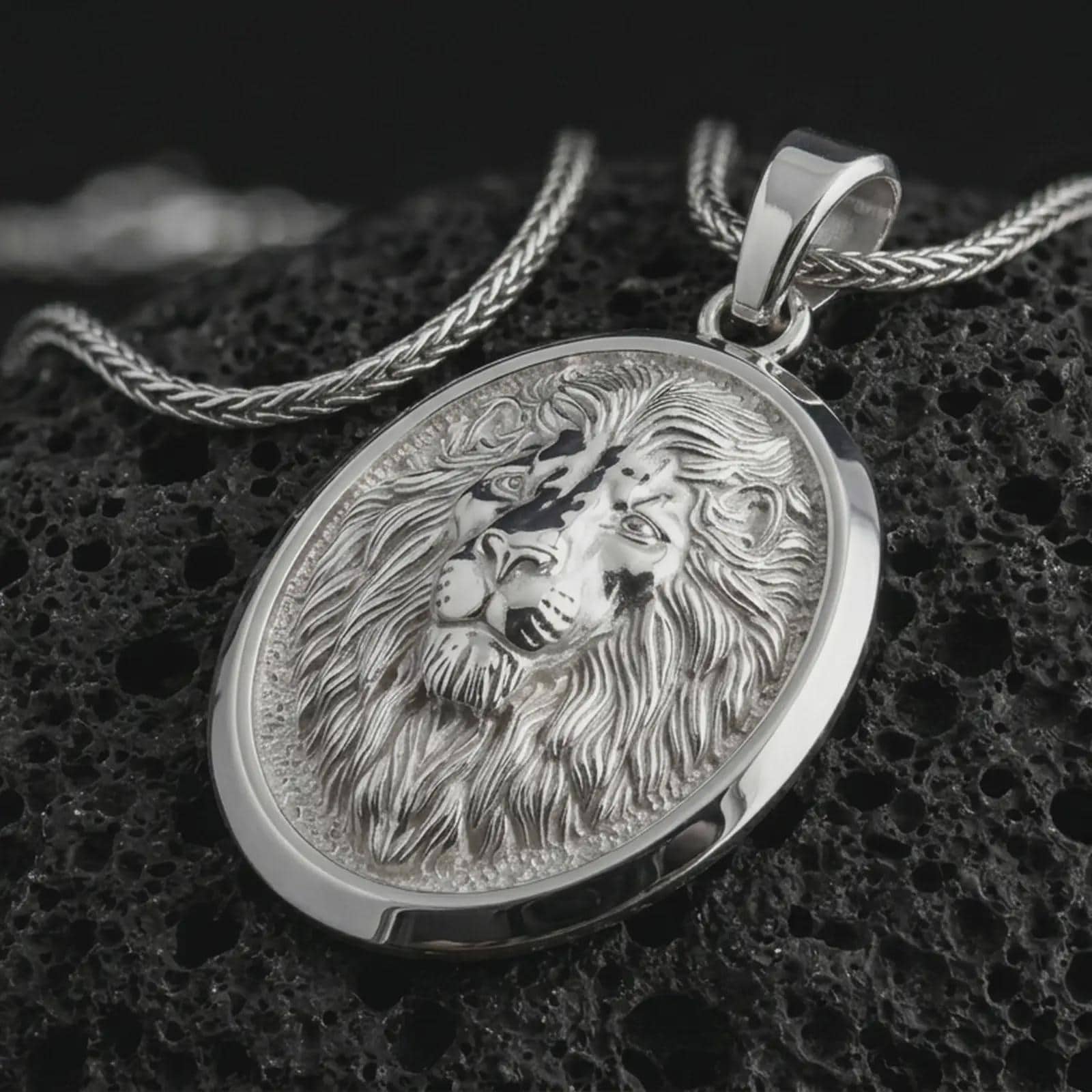 Polished silver lion medallion pendant necklace displayed on black volcanic rock, shown in a refined macro product shot with majestic lion face relief, flowing mane detail, and oval frame.