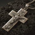 Load image into Gallery viewer, Bronze Jesus Ascension cross pendant necklace with an oxidized finish resting on textured volcanic rock, captured in a close-up premium studio composition.
