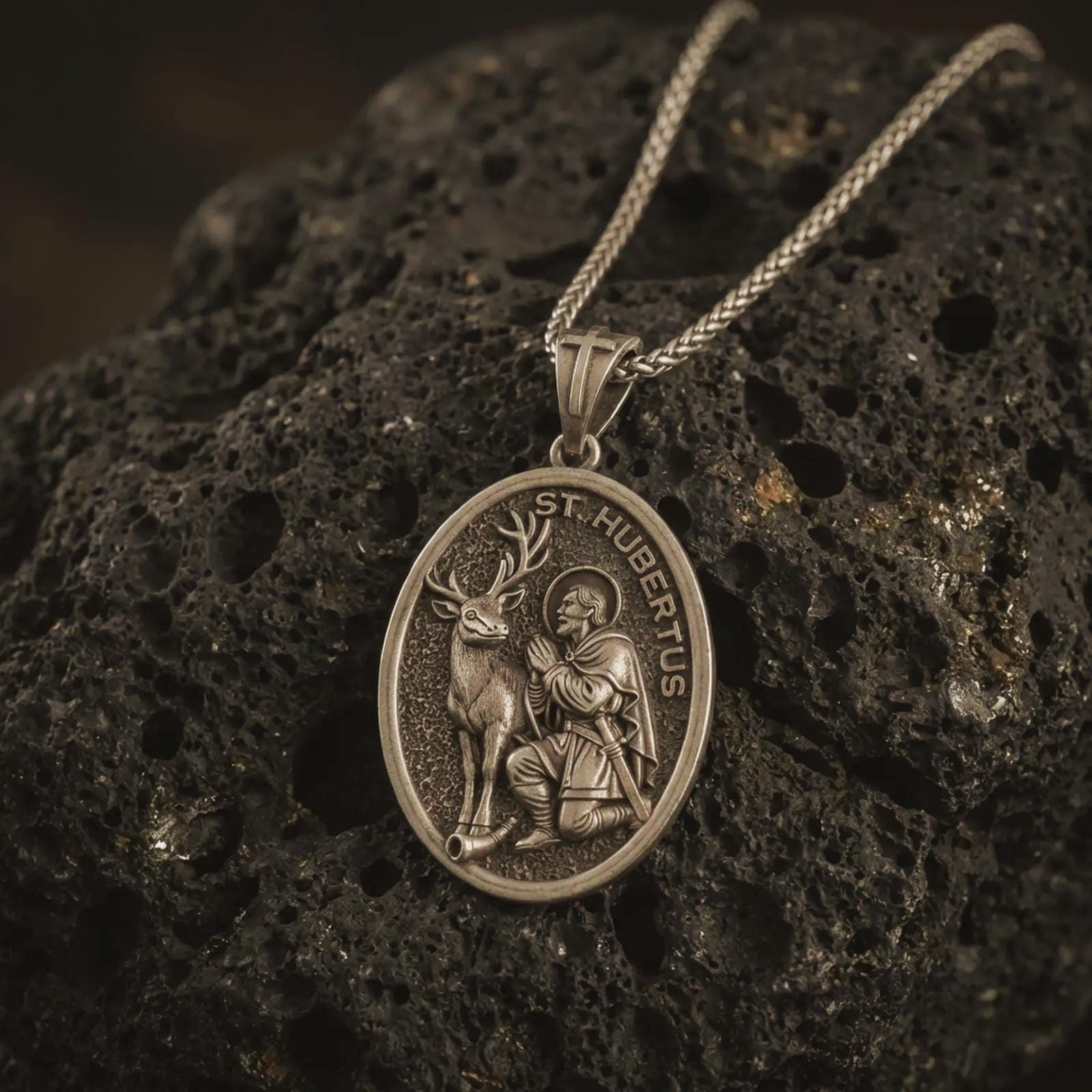 Bronze Saint Hubert pendant necklace with an oxidized finish resting on textured volcanic rock, captured in a close-up premium studio composition.