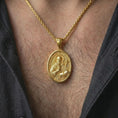 Load image into Gallery viewer, Gold plated Saint Benedict pendant necklace worn on the neck, captured on a human model in a premium dark-toned lifestyle frame.
