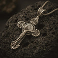 Load image into Gallery viewer, Bronze Saint Benedict crucifix necklace with an oxidized finish resting on textured volcanic rock, captured in a close-up premium studio composition.
