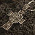 Load image into Gallery viewer, Bronze Orthodox cross pendant necklace with an oxidized finish resting on textured volcanic rock, captured in a close-up premium studio composition.
