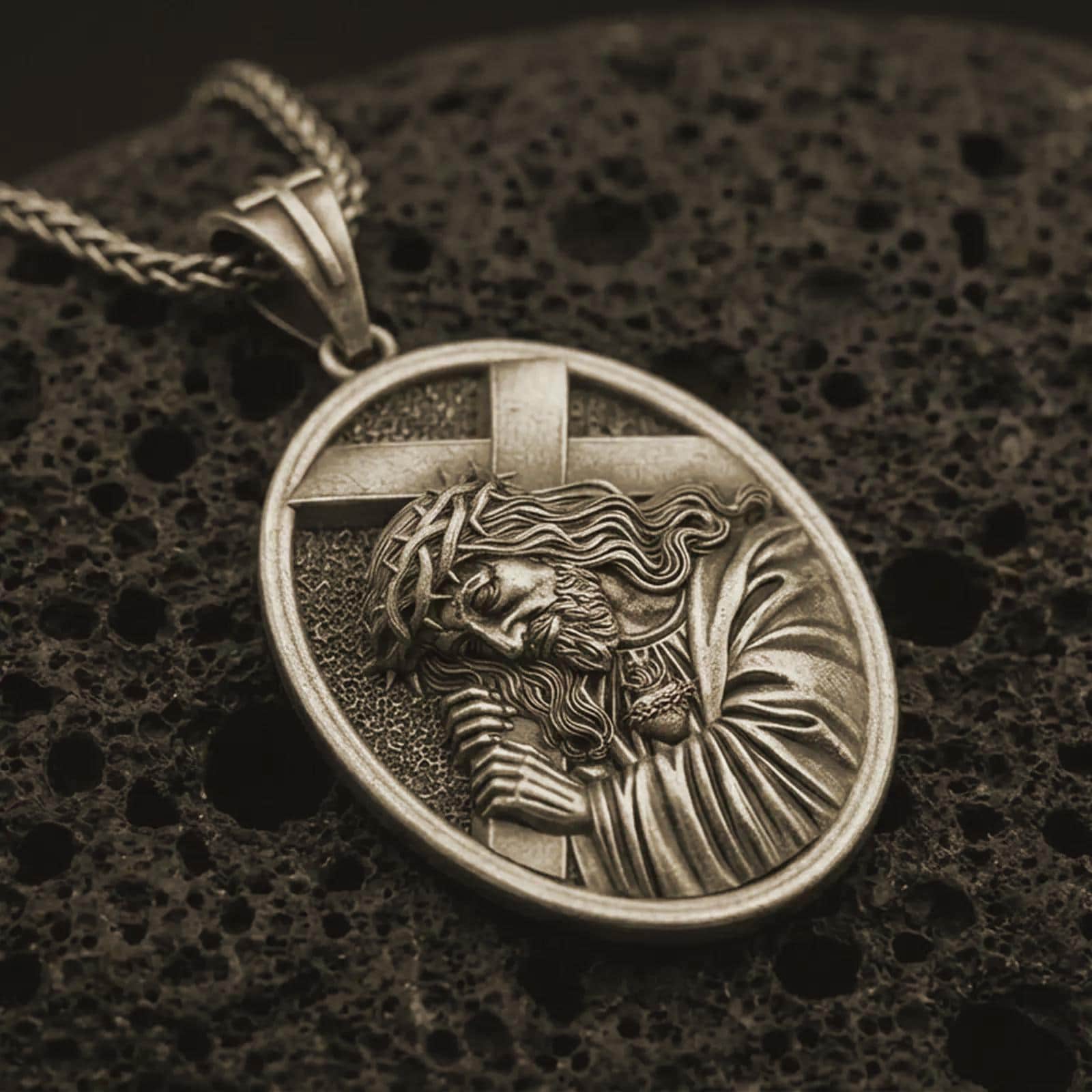 Bronze Jesus carrying cross pendant necklace with an oxidized finish resting on textured volcanic rock, captured in a close-up premium studio composition.