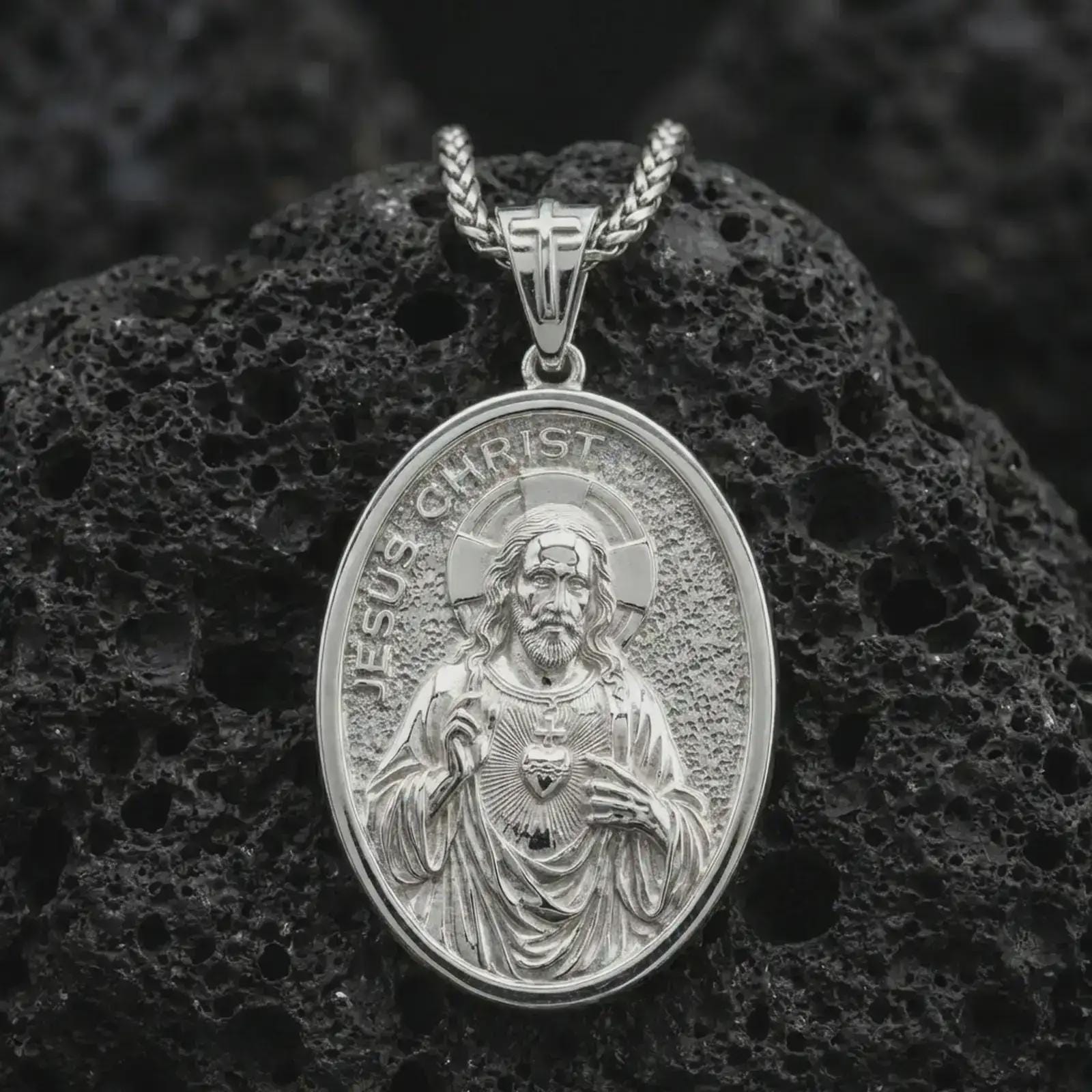 Polished silver Jesus Christ pendant necklace displayed on black volcanic rock, shown in a refined macro product shot with detailed sacred figure and engraved lettering.