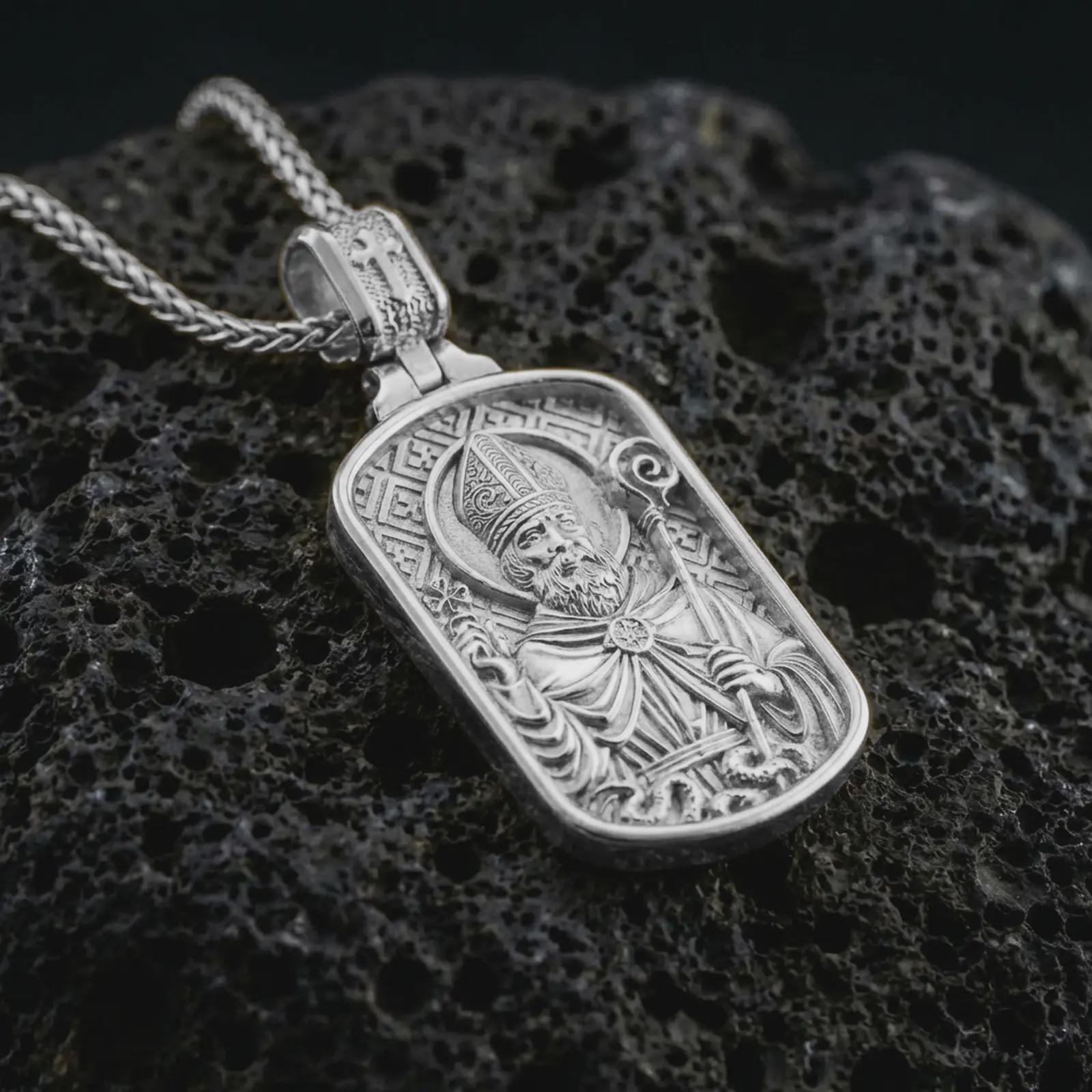 Polished silver Saint Patrick dog tag pendant necklace displayed on black volcanic rock, shown in a refined macro product shot with detailed bishop figure, staff, and engraved border pattern.