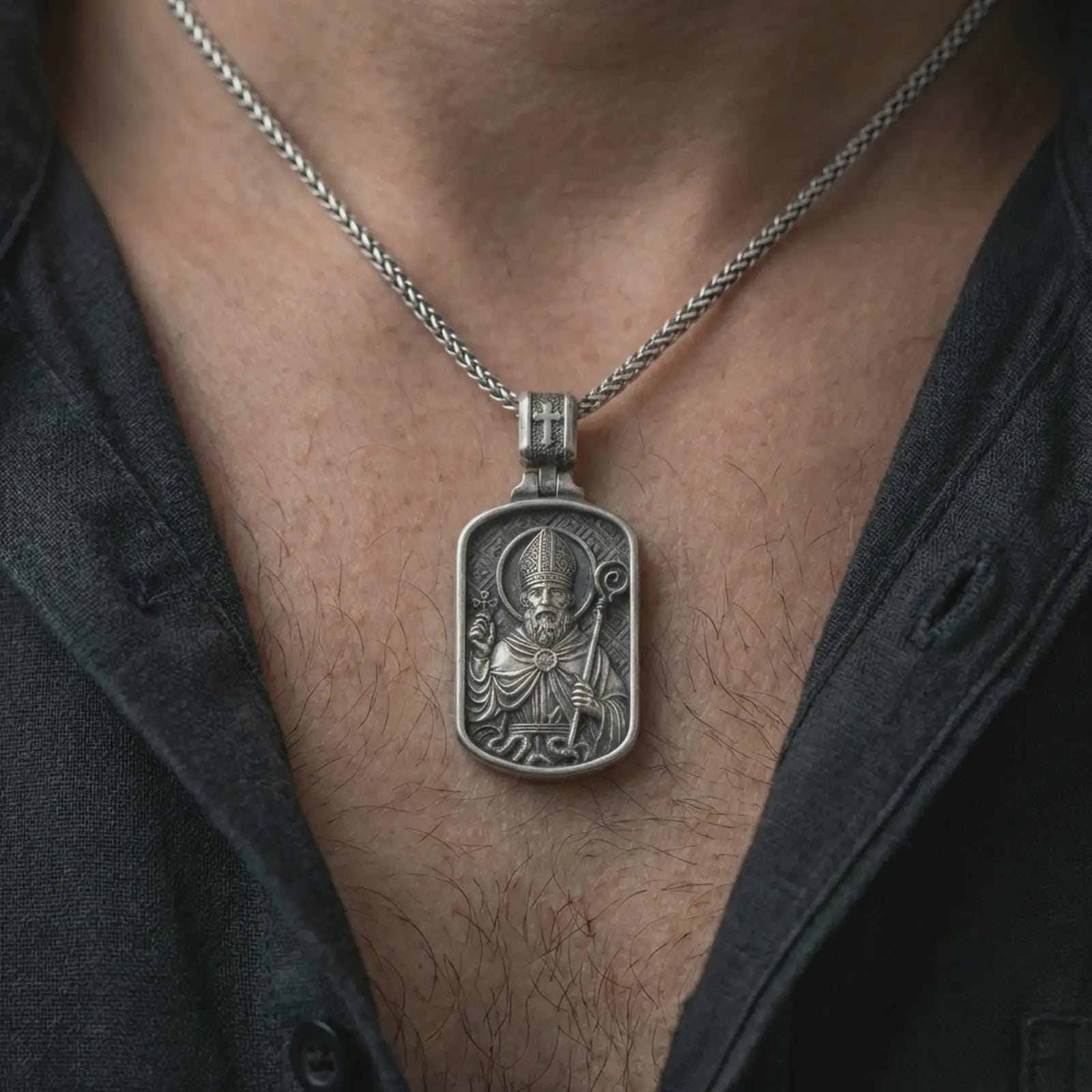 Oxidized Saint Patrick dog tag pendant necklace worn on the neck, styled in a dark lifestyle portrait with an open shirt and masculine presentation.