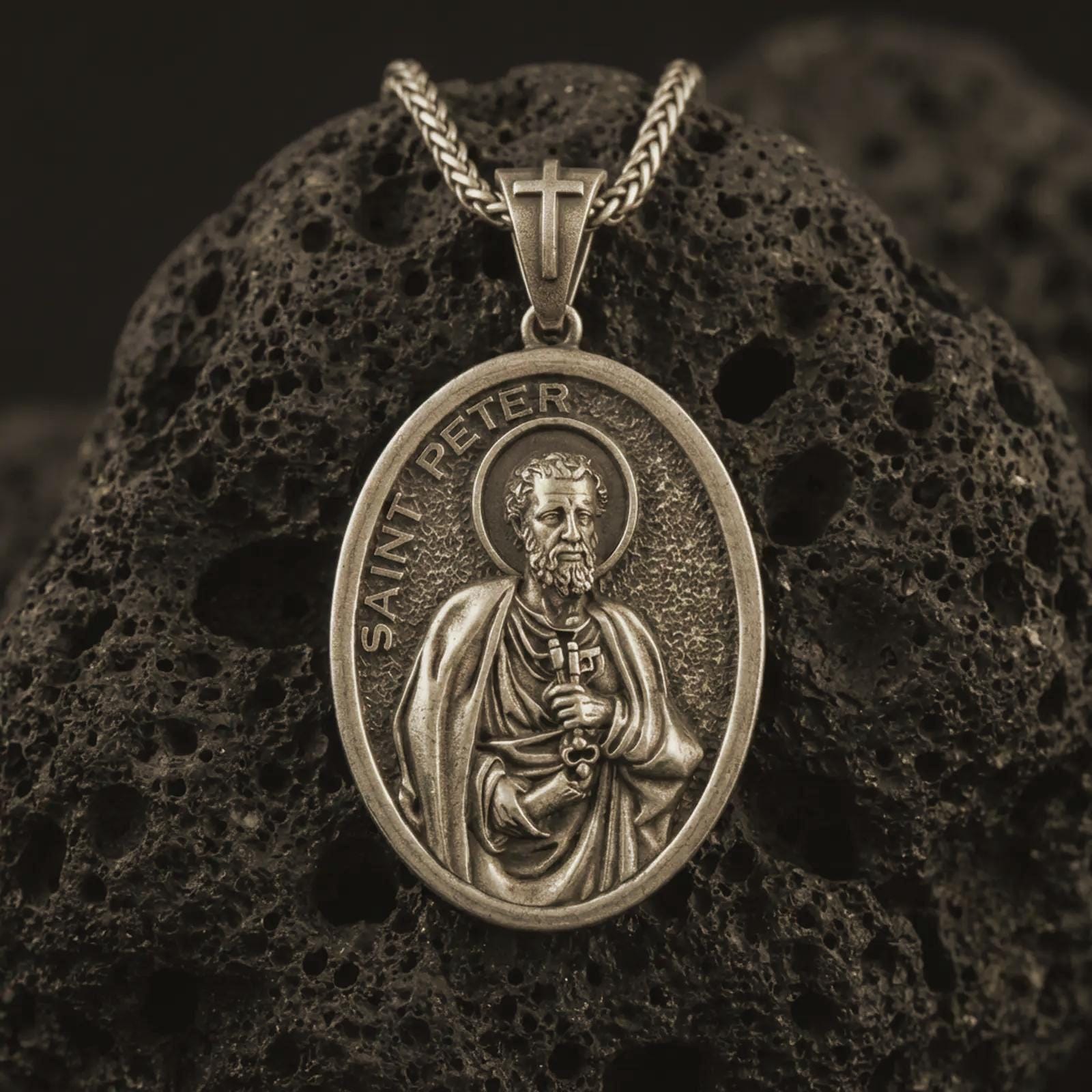 Bronze Saint Peter pendant necklace with an oxidized finish resting on textured volcanic rock, captured in a close-up premium studio composition.