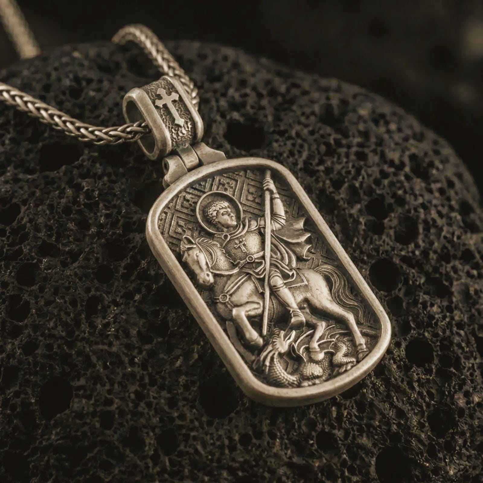 Bronze Saint George dog tag pendant necklace with an oxidized finish resting on textured volcanic rock, captured in a close-up premium studio composition.