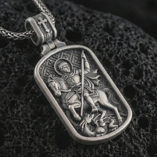 Sterling silver Saint George dog tag pendant necklace with an oxidized finish resting on textured volcanic rock, captured in a close-up premium studio composition.