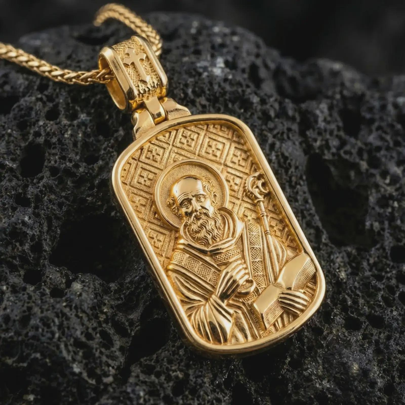 Gold plated Saint Benedict dog tag pendant necklace presented on porous volcanic rock, highlighting the rectangular devotional form in a luxurious close-up jewelry image.