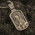 Load image into Gallery viewer, Bronze Saint Benedict dog tag pendant necklace with an oxidized finish resting on textured volcanic rock, captured in a close-up premium studio composition.
