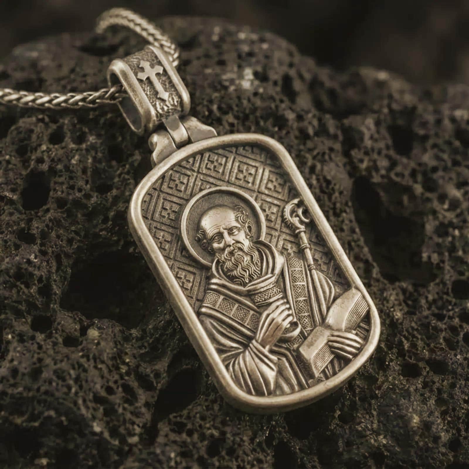 Bronze Saint Benedict dog tag pendant necklace with an oxidized finish resting on textured volcanic rock, captured in a close-up premium studio composition.