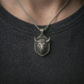 Load image into Gallery viewer, Oxidized bull head shield pendant necklace worn on the neck, styled in a dark lifestyle portrait with an open shirt and bold masculine presentation.

