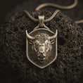 Load image into Gallery viewer, Bronze bull head shield pendant necklace with an oxidized finish resting on textured volcanic rock, captured in a close-up premium studio composition.

