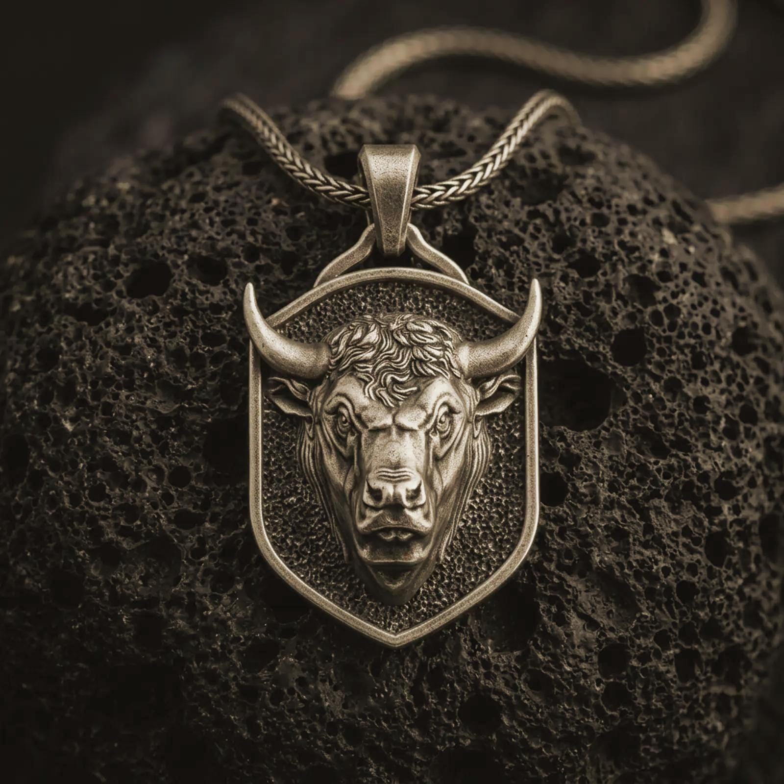 Bronze bull head shield pendant necklace with an oxidized finish resting on textured volcanic rock, captured in a close-up premium studio composition.