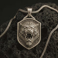 Load image into Gallery viewer, Bronze roaring lion shield pendant necklace with an oxidized finish resting on textured volcanic rock, captured in a close-up premium studio composition.
