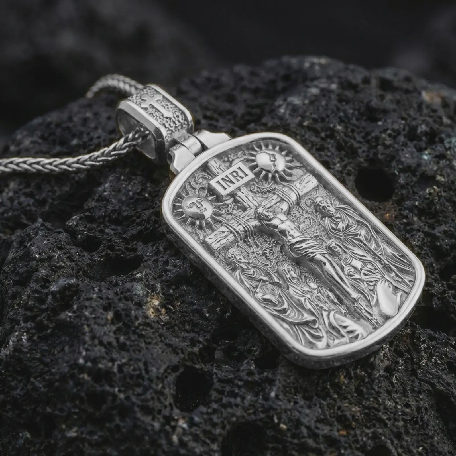 Polished silver Jesus INRI crucifixion pendant necklace displayed on black volcanic rock, shown in a refined macro product shot with detailed crucifix scene and sacred figure relief.