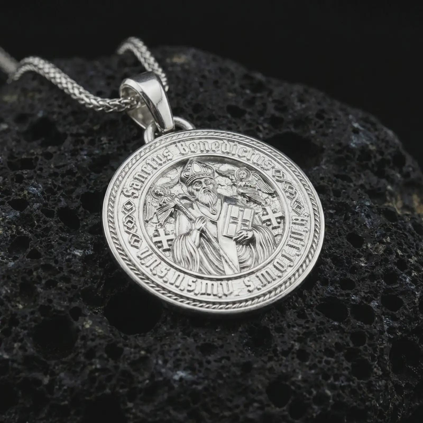 Polished silver Saint Benedict medal pendant necklace displayed on black volcanic rock, shown in a refined macro product shot with detailed saint figure and engraved Latin lettering.