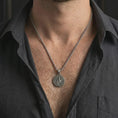 Load image into Gallery viewer, Oxidized Saint Benedict medal pendant necklace worn on the neck, styled in a dark lifestyle portrait with an open shirt and masculine presentation.
