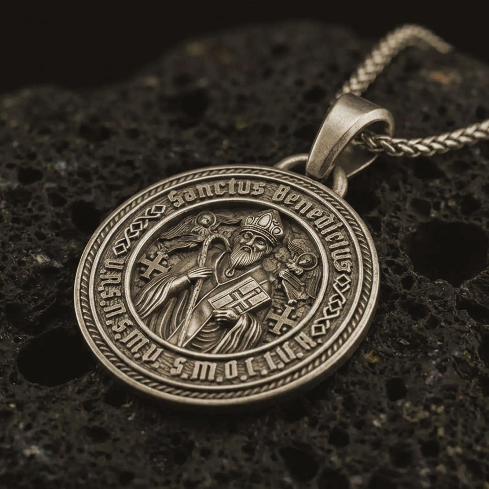 Bronze Saint Benedict medal pendant necklace with an oxidized finish resting on textured volcanic rock, captured in a close-up premium studio composition.