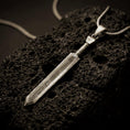 Load image into Gallery viewer, Bronze Dragonslayer sword pendant necklace with an oxidized finish resting on textured volcanic rock, captured in a close-up premium studio composition.
