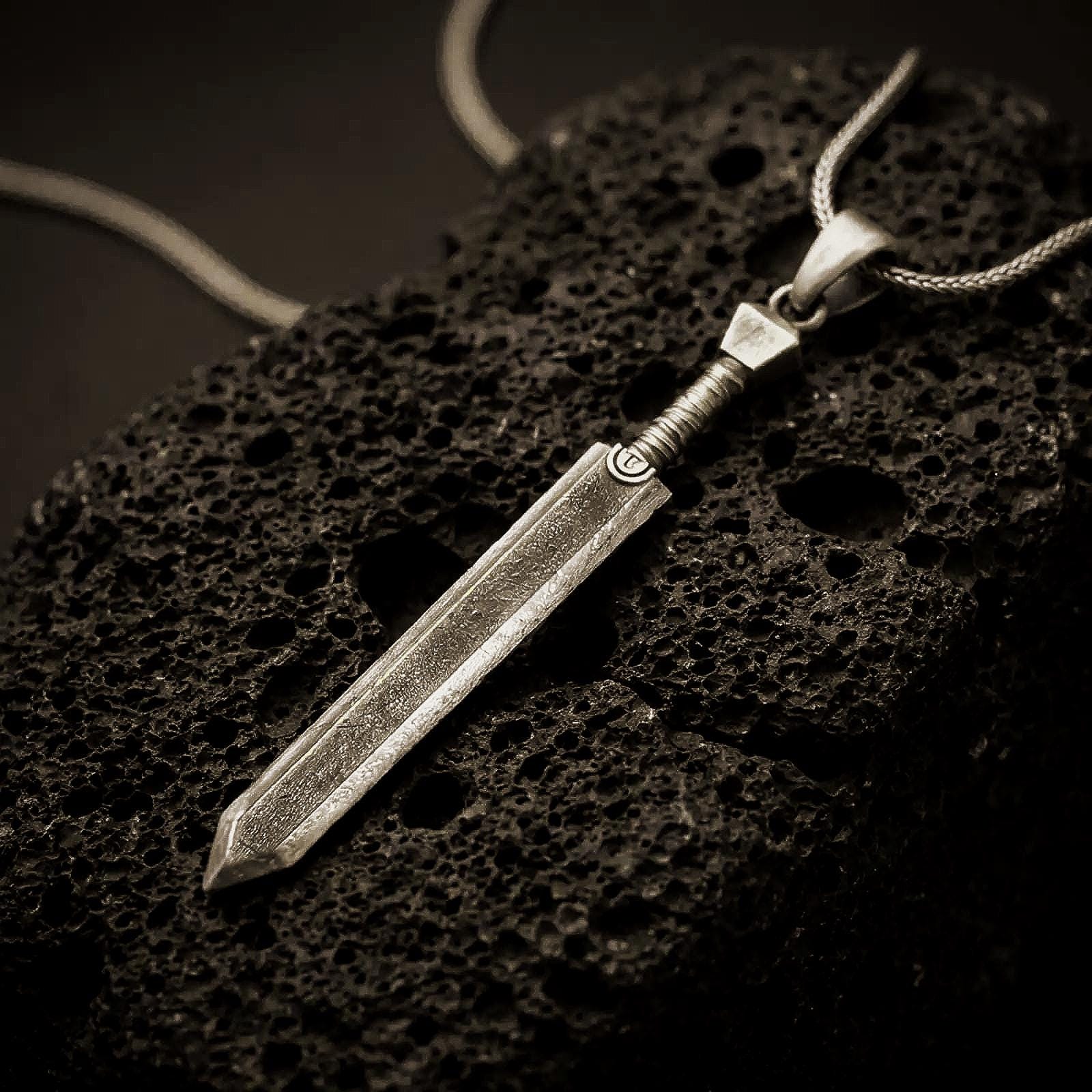 Bronze Dragonslayer sword pendant necklace with an oxidized finish resting on textured volcanic rock, captured in a close-up premium studio composition.