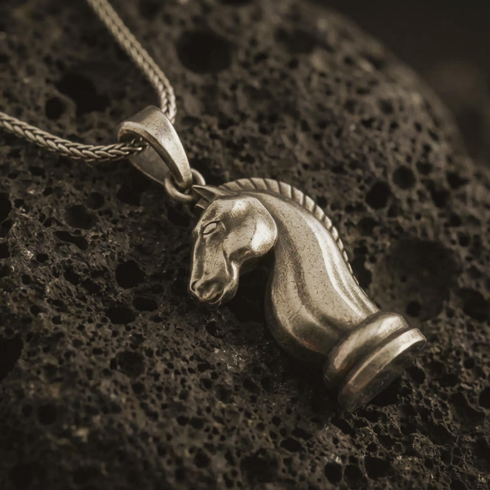 Bronze knight chess piece pendant necklace with an oxidized finish resting on textured volcanic rock, captured in a close-up premium studio composition.