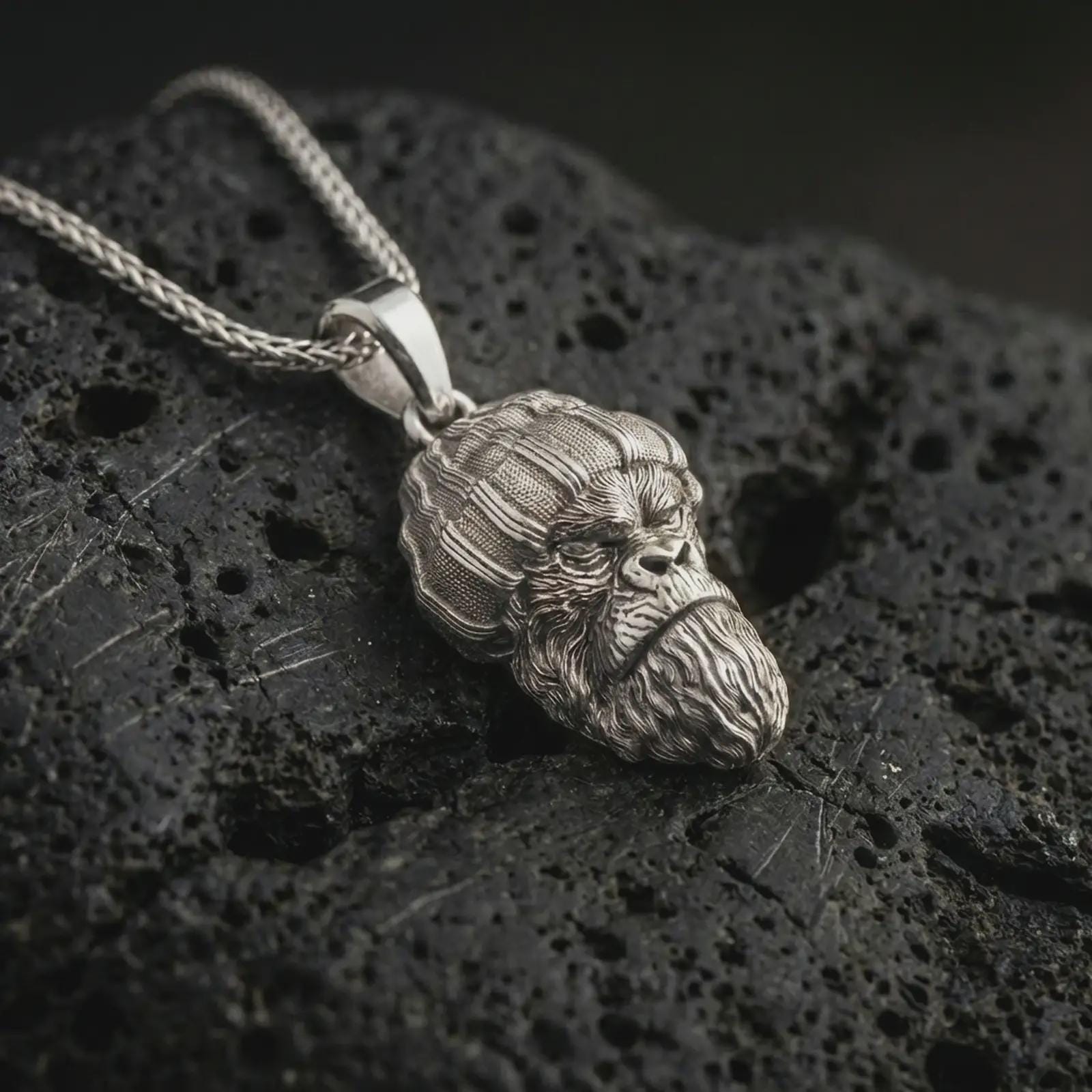 Polished silver gorilla head pendant necklace displayed on black volcanic rock, shown in a refined macro product shot with knit beanie detail, sculpted beard texture, and urban primate character.