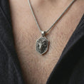 Load image into Gallery viewer, Oxidized ram head shield pendant necklace worn on the neck, styled in a dark lifestyle portrait with an open shirt and bold Aries-inspired presentation.
